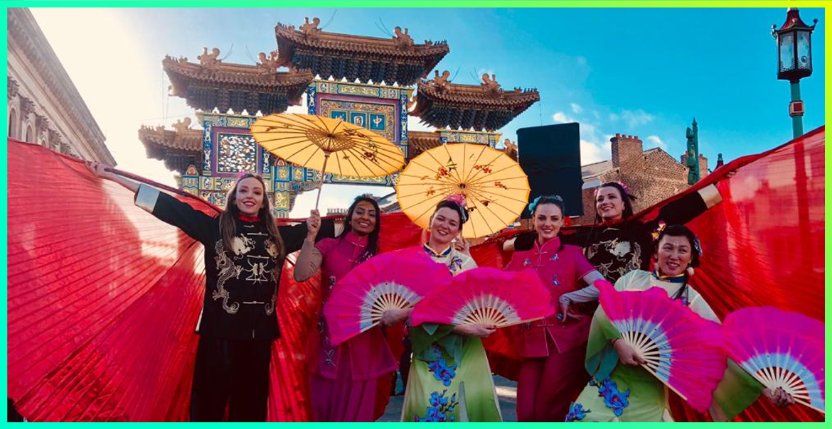 a group of woman standing in front of the Chinese Arch in Liverpool, dressed in mandarin clothing holding parasols and fans