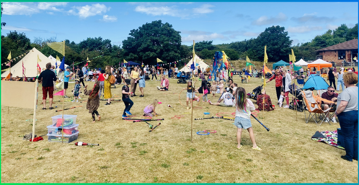 A family-friendly festival happening in a field on a sunny day with children playing, colourful bunting and yurts.
