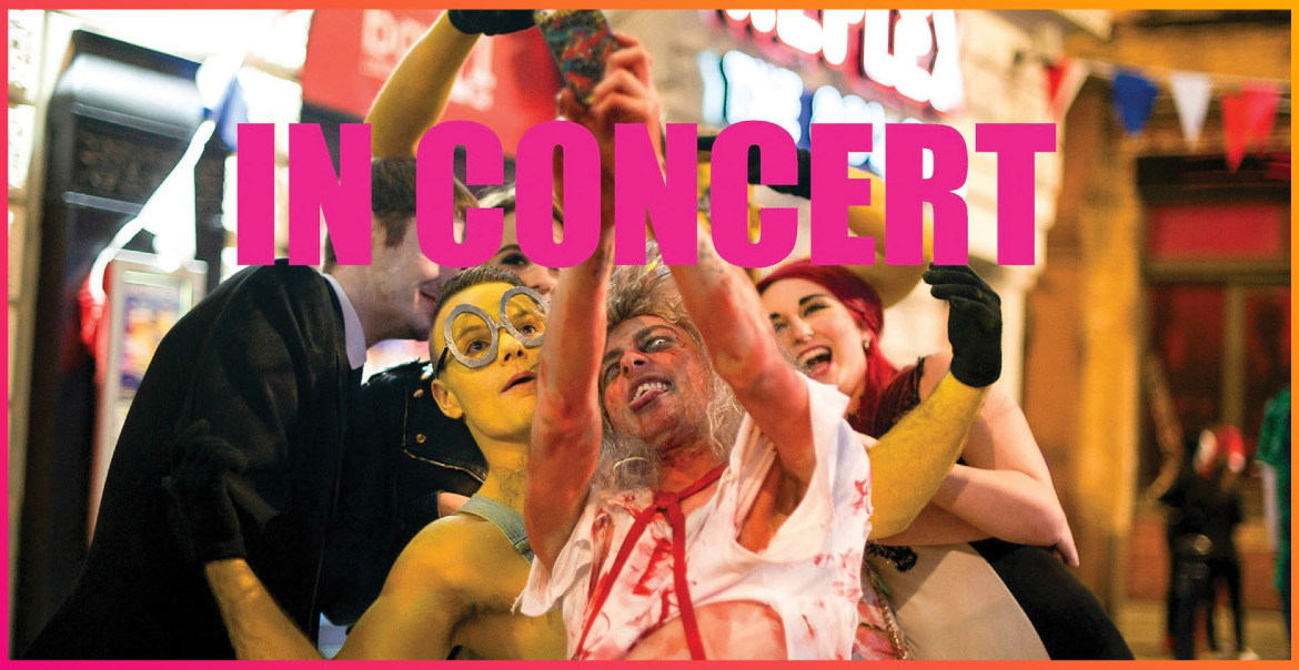 A group of people dressed up for Halloween taking a selfie together. Pink text reads "IN CONCERT" over the image.