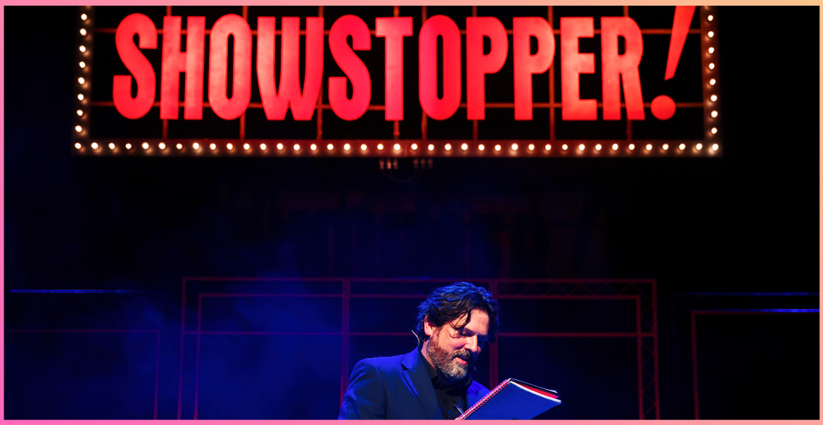 Production image of 'Showstopper' featuring bright red lights spelling out the title and a male character on stage reading out of a book.