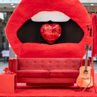 A large red mouth sculpture with a red mirror ball in between the teeth. In front of the mouth is a red couch, red-glittered guitar and more red items like a phone.