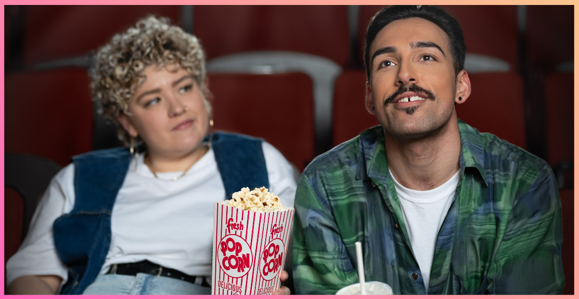 A man and woman sitting in a cinema auditorium with popcorn and a drink. The woman looks at the man while the man watches the screen.