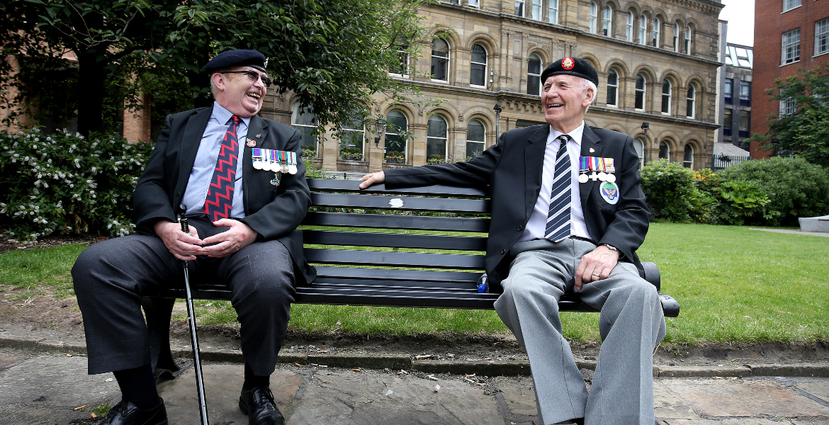 two men sat on a bench in smiling at each other wearing their military medals