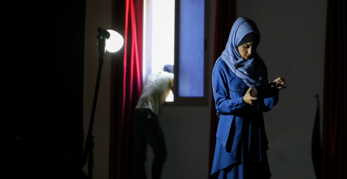 A woman looking down in a dark room while a man behnd her leans out the window.