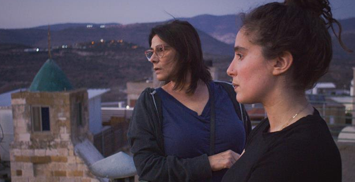 Two women stand on a rooftop overlooking a mountainous area at dusk, gazing into the distance. The older woman has glasses and short hair; the younger has her hair tied back.