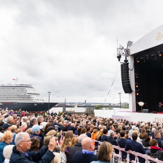 A large crowd over looking the Queen Anne cruise ship on an overcast day. A large, outdoor stage stands in front of the crowd with Cunard branding.