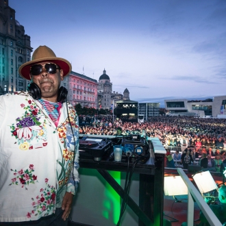 A DJ facing the camera at an outdoor event with the crowd and Liveprool iconic buildings in the background.
