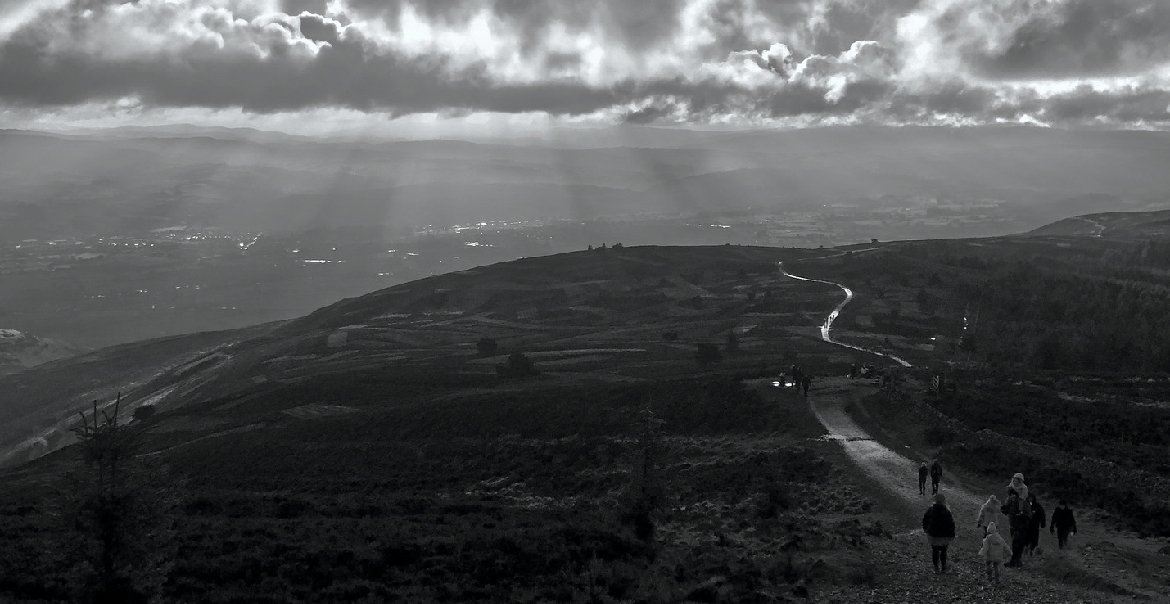 A black and white image of hills with the sun shining through the clouds.