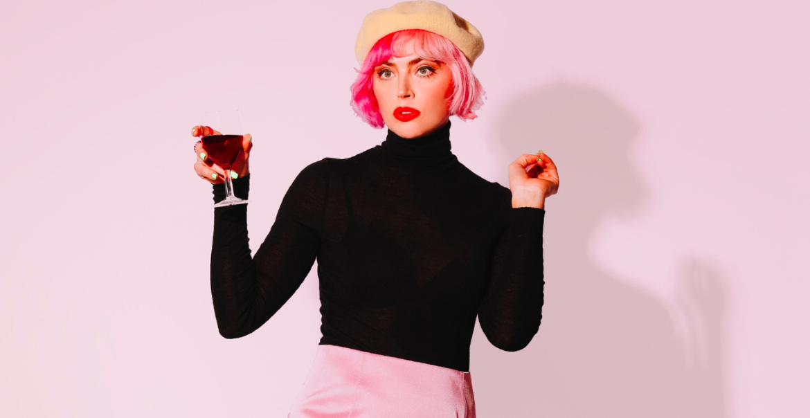 An image of a woman wearing a long-sleeved black top, a cream beret and holding out a drink.