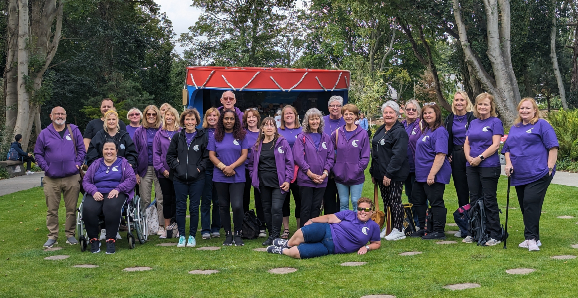 A large choir standing in front of a bandstand in a park. The choir members are wearing purple t-shirts.