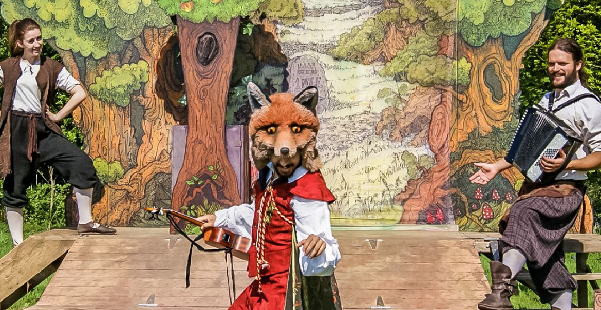 A production image of an outdoor performance featuring two actors and a persond wearing a fox costume