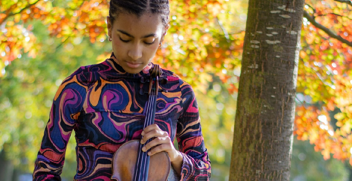 A woman holding a violin in the middle of an outdoor space with trees.