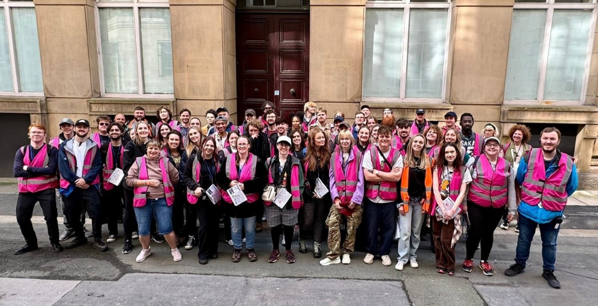 A diverse group of people wearing pink vests smiling together in front of a large building, showcasing teamwork.