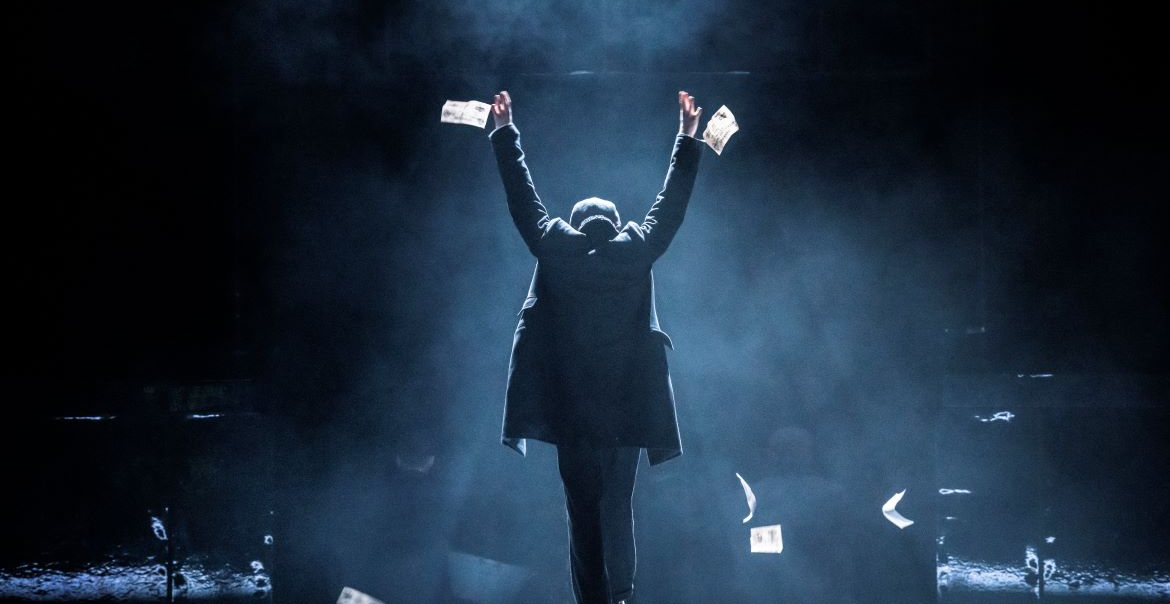 A prdouction image of a Peaky Blinders theatre show featuring the character Tommy Shelby, wearing a flat cap and a long coat, with his back to the audience and his hands in the air.