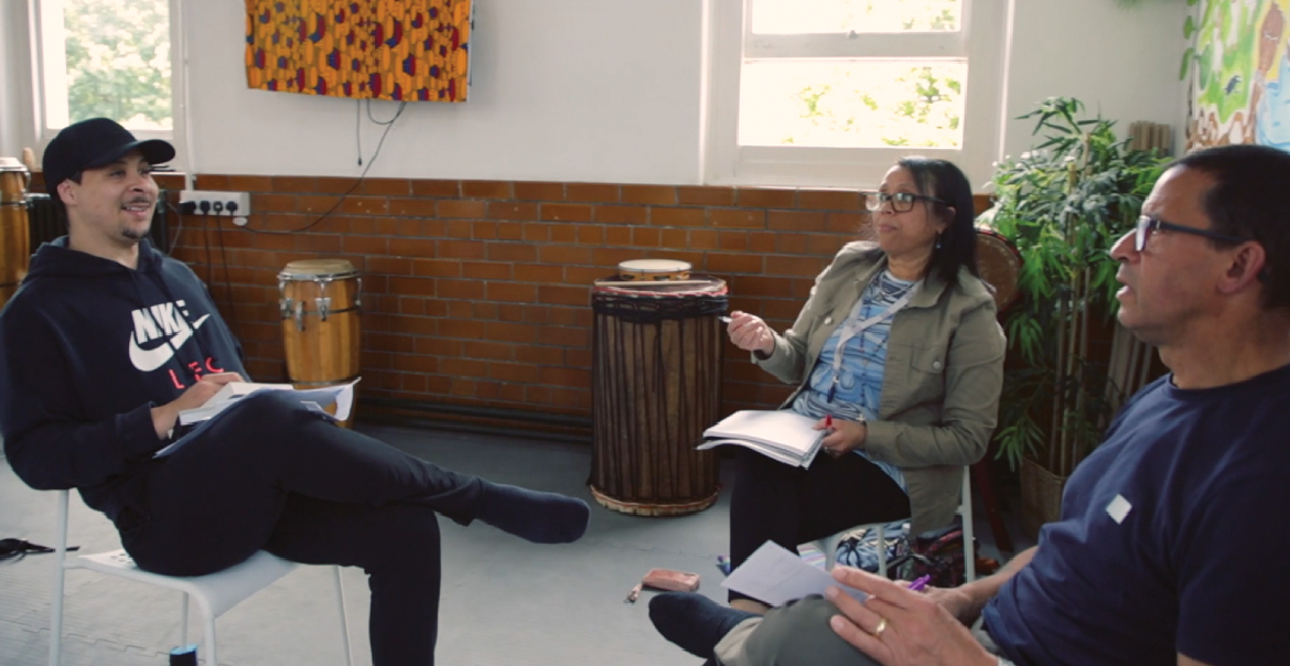 Three people engaged in conversation, each sat on a chair in a community space.