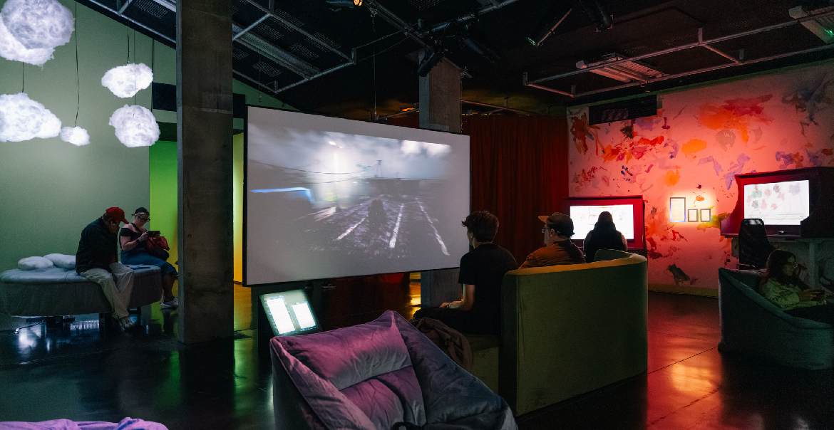 Visitors are seated on bean bags and sofas in a dimly lit, atmospheric room at an art exhibition. The walls are illuminated with colorful light projections, and fluffy cloud-like installations hang from the ceiling. A large screen displays a video game.