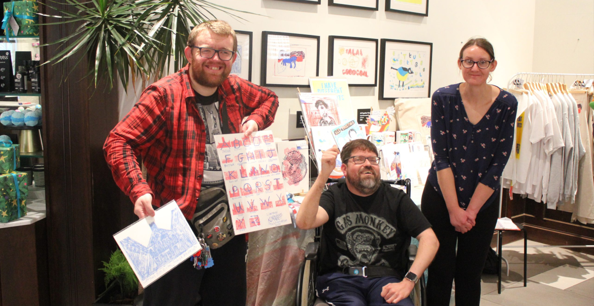 Three individuals posing in a store with artwork. One is seated in a wheelchair, and the others are standing on either side, all holding framed illustrations. They are smiling and surrounded by various clothing items on display.