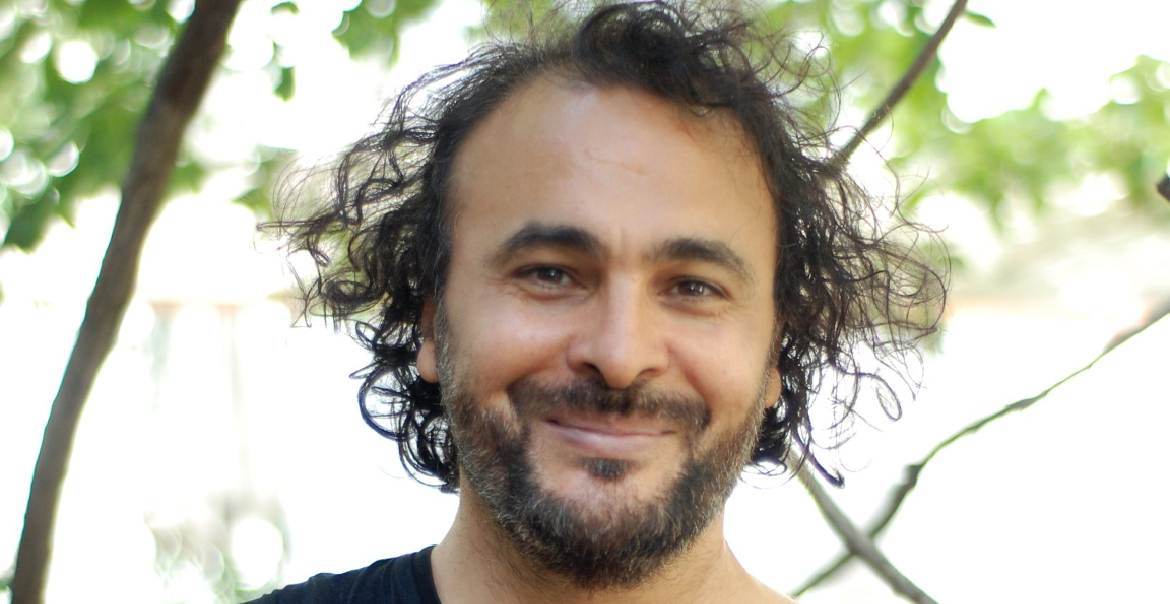 A portait image of a man who is standing outstide on a bright day,. Trees can be seen in the background.