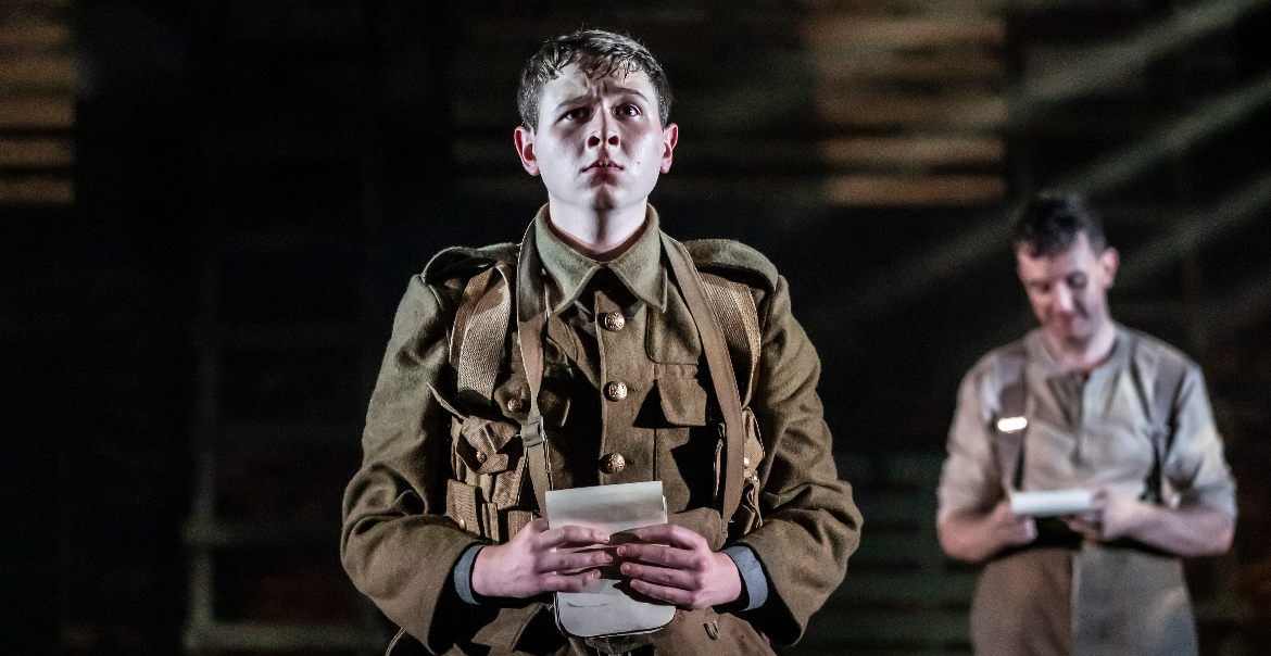 A production image from the stage play, Birdsong, featuring a soldier holding a letter and staring out towards the audience while a character stand behind them,