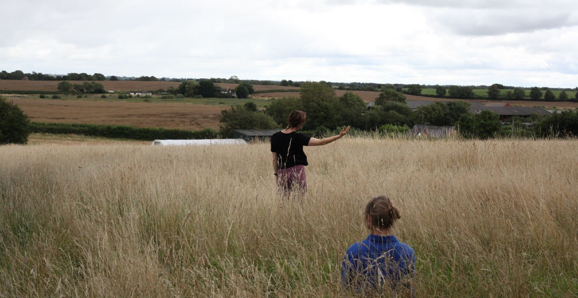 An image of two people in a rural field on a cloudy day. One is sat down and one, a little further ahead, stands with the right arm raised to the side.