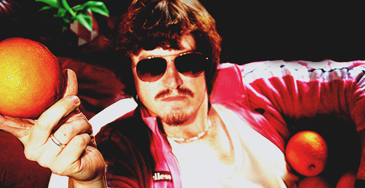 A film photograph of a mn wearing a pink jacket, sunglasses and holding up an orange.