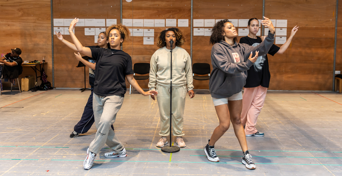 Four dancers practice a choreographed routine in a studio with a wooden floor and a wall covered with notes in the background.