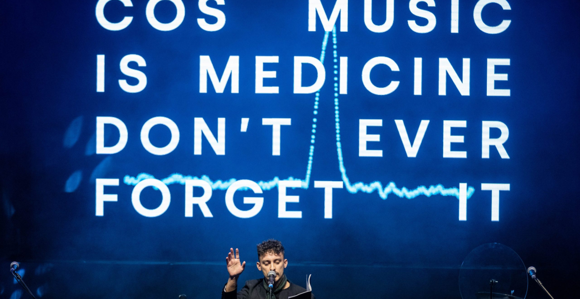 A person perfomring on stage with words projected behind them reading 'COS MUSIC IS MEDICINE DON'T EVER FORGET IT'